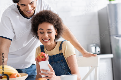 cheerful interracial couple looking at smartphone during breakfast