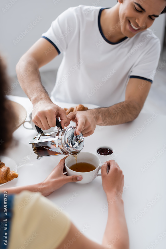 happy man pouring tea in cup near blurred african american girlfriend