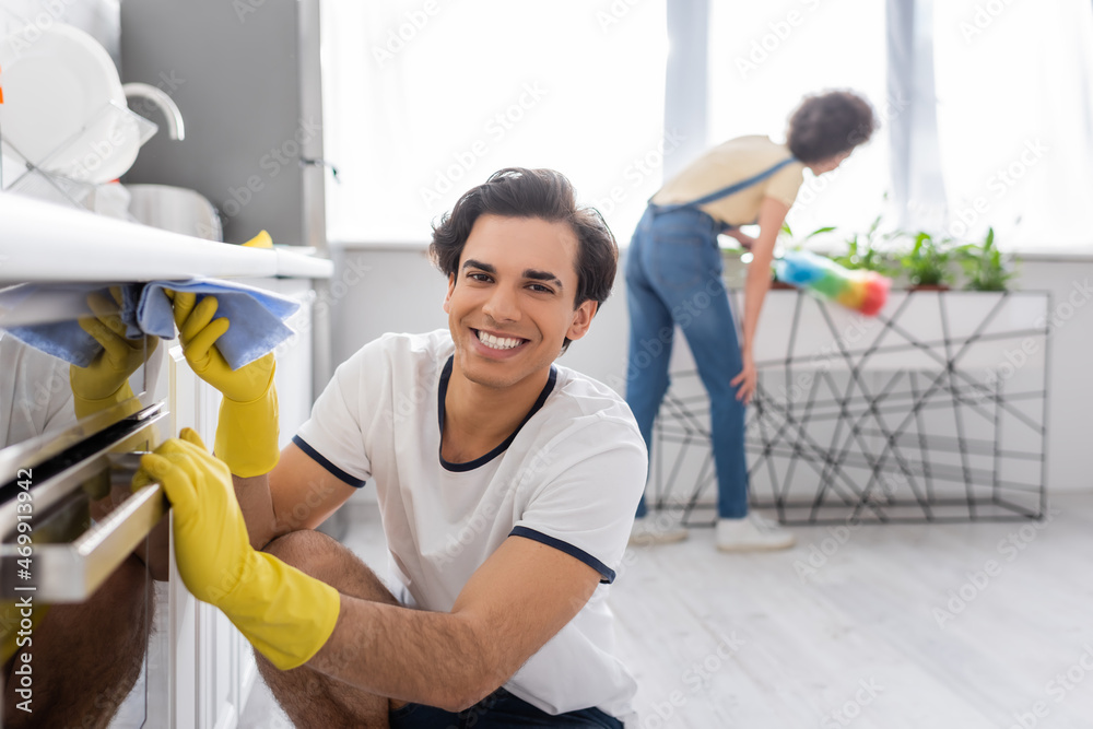 Obraz premium happy young man cleaning electric oven with rag near curly african american woman with dust brush in kitchen