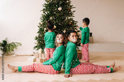 The concept of Christmas. Cheerful children in green pajamas have fun tied up their parents with a rope and stole gift boxes from under the Christmas tree during the New Year holidays at home.