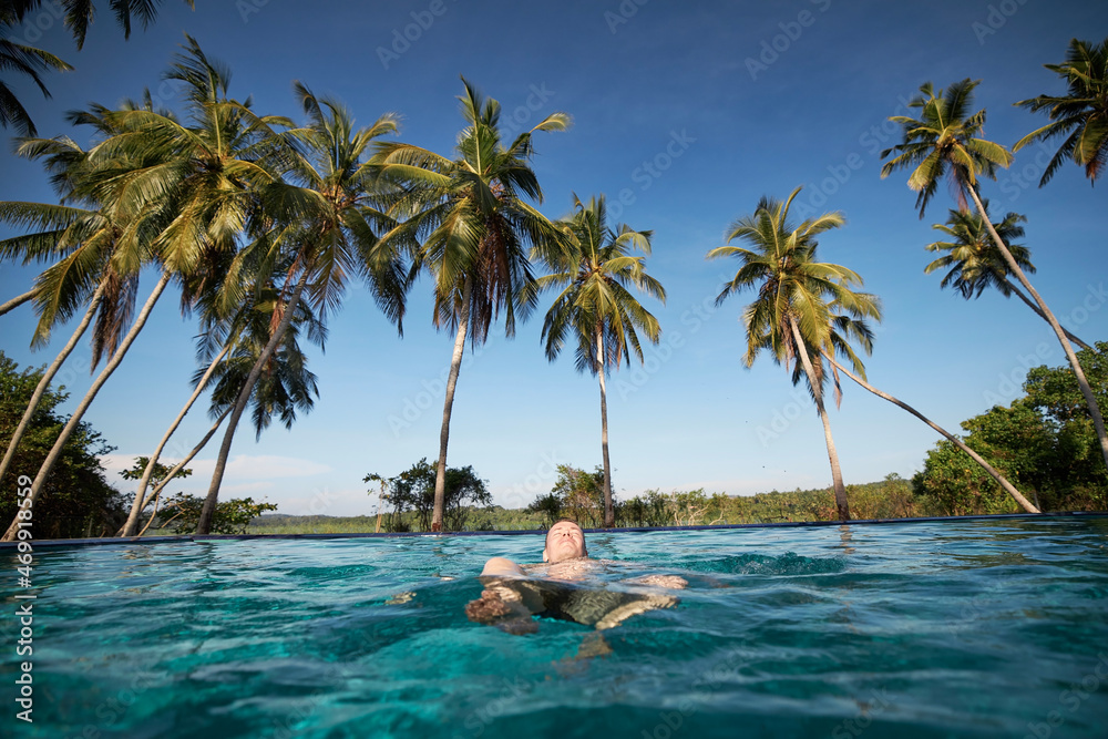 Man is swimming on back in pool against palm trees. Relaxation in the middle of beautiful tropical nature in Sri Lanka..