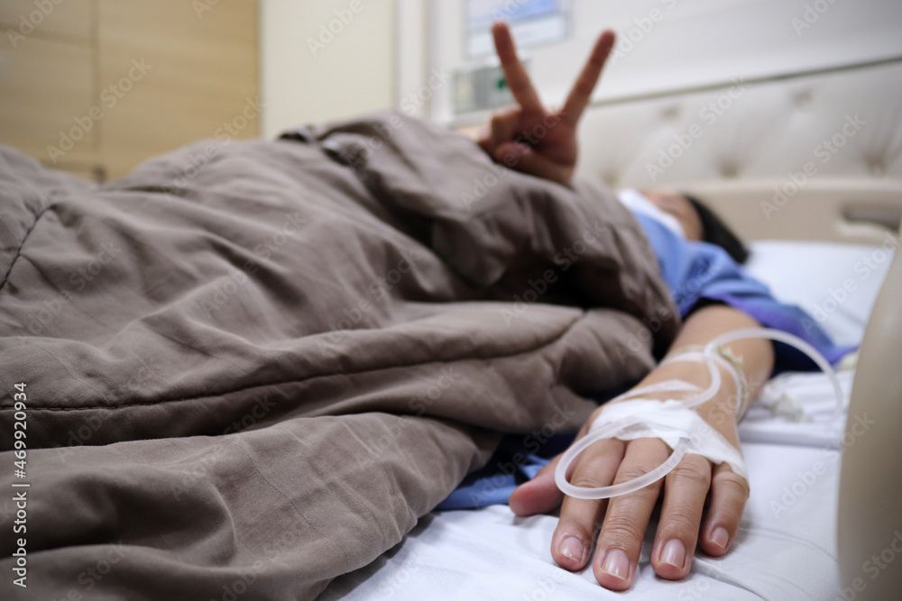 Asian woman wearing medical hygiene face mask showing blurry finger in morale sign with happy and focus on the hand of patient in hospital ward