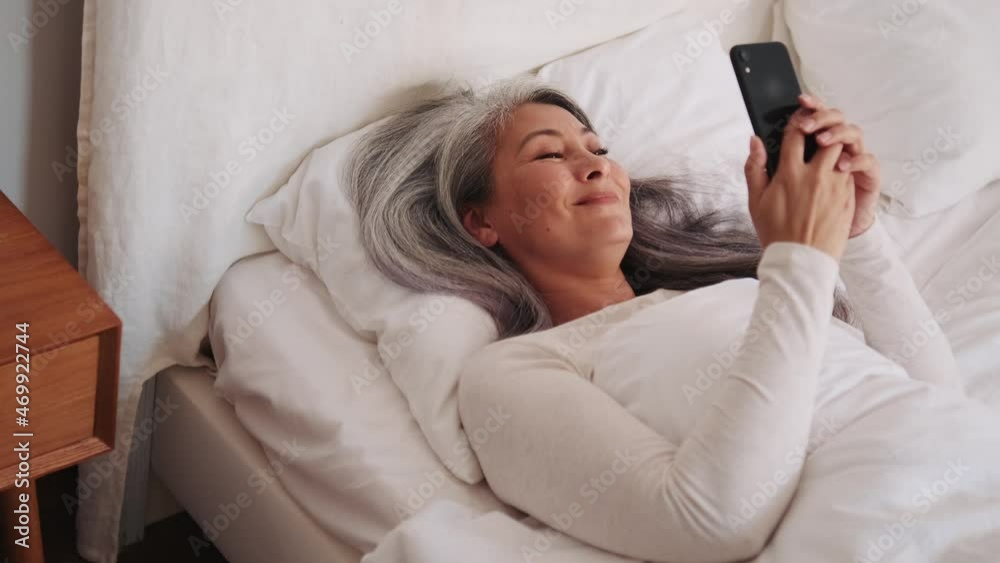 Positive Asian woman typing by phone in the bed Positive Asian woman typing by phone in the bed