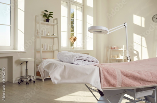 Beautiful interior of empty spa salon, massage room, beauty centre, or dermatologist's office with massage table, modern lamp magnifier, white walls, and shelves with products for skin care procedures photo