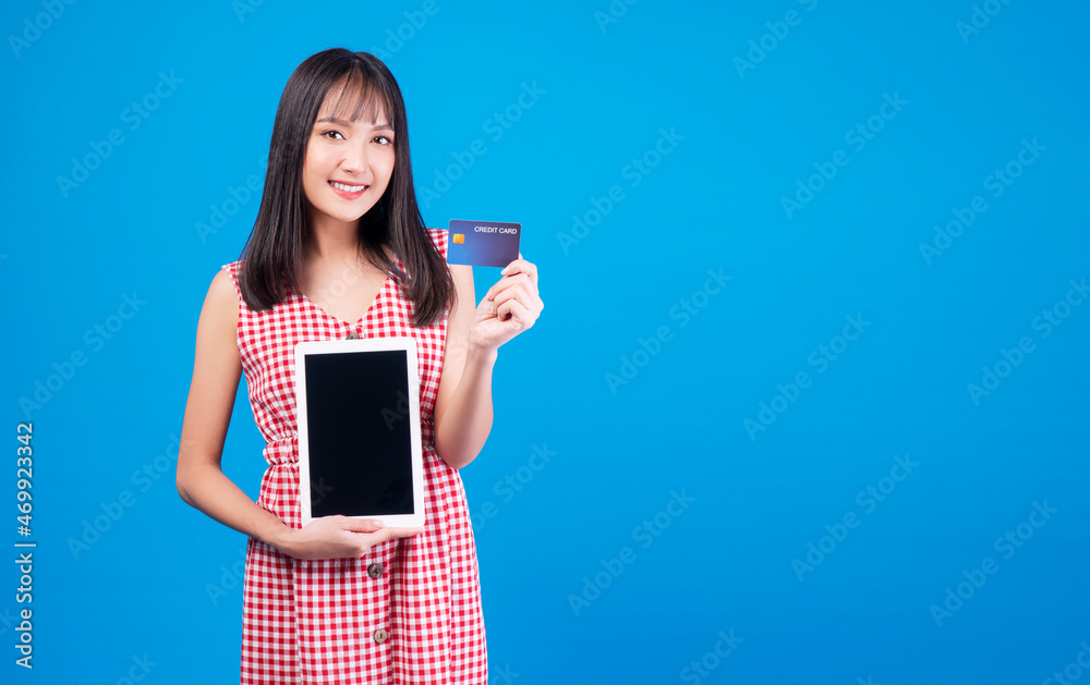 Financial payment online shopping and money transfer concept. Excited smile young woman holding digital tablet and mockup credit card for internet banking while standing over isolated blue background. Financial payment online shopping and money transfer concept. Excited smile young woman holding digital tablet and mockup credit card for internet banking while standing over isolated blue background.