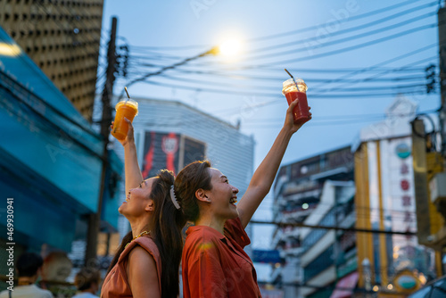 Asian woman friends walking and shopping together at chinatown street night market in Bangkok city, Thailand. Female tourist enjoy outdoor lifestyle travel drink fruit juice and eating street food.