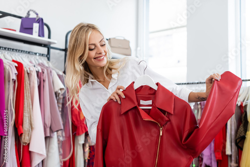 happy tattooed woman holding red leather jacket on hanger while choosing clothes