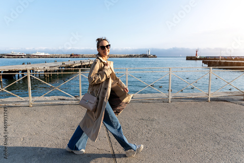 Yonug adult trendy stylish beautiful caucasian happy smiling woman enjoy walking by Yalta sea embankment on warm sunny day. Female person portrait wear jeans biege trench coat on urban city street