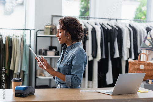 cheerful african american saleswoman holding digital tablet near gadgets and paper cup on sales counter desk photo