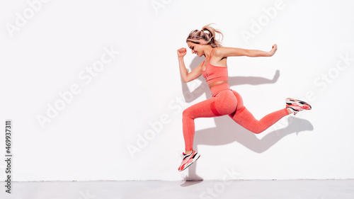Fitness smiling woman in pink sports clothing. Young beautiful model with perfect body. Female posing in the street near white wall at summer sunny day. Cheerful and happy. Jumping and running
