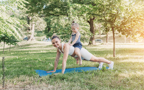 The baby is sitting on the back of her mother, who is doing exercises on a yoga mat in the park on the grass.