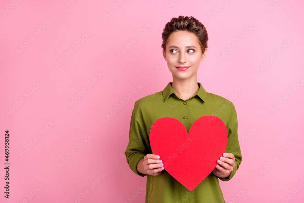 Photo of adorable tricky young woman dressed green shirt smiling holding red heart looking empty space isolated pink color background Photo of adorable tricky young woman dressed green shirt smiling holding red heart looking empty space isolated pink color background