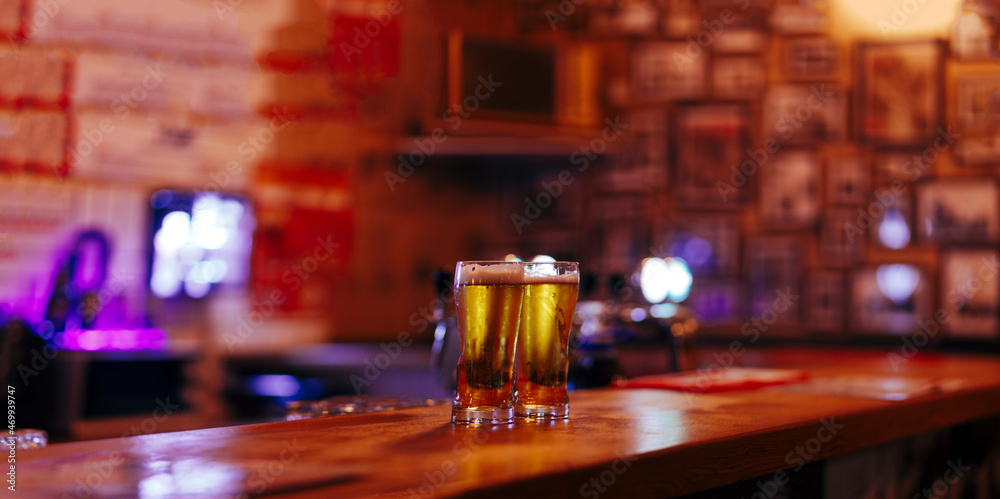 two glasses full of cold beer against the blurry empty interior of a beer bar