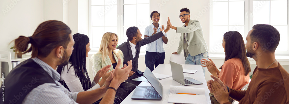 Mixed race business team celebrating good results in office meeting. Happy young black man gives high five to coworker while diverse multi ethnic teammates are applauding. Teamwork and success concept - obrazy, fototapety, plakaty 