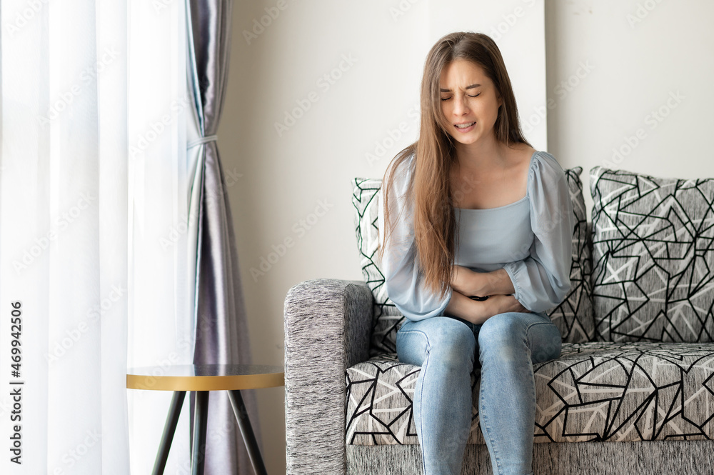 Young woman suffering from strong stomach pain while sitting on sofa at home. health problems Young woman suffering from strong stomach pain while sitting on sofa at home. health problems