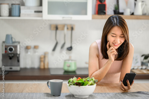 Happy smiling Asian woman using smartphone mobile at home in kitchen with salad bowl on the table
