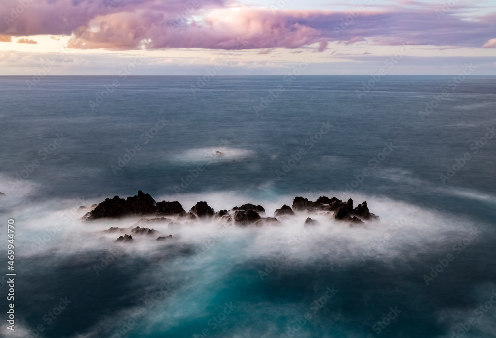 Porto Moniz on the north coast of Madeira island Portugal with dark lava rocks and islands near shore. Dusk sunset seascape panorama with and turquoise water and colorful clouds, longtime exposure.