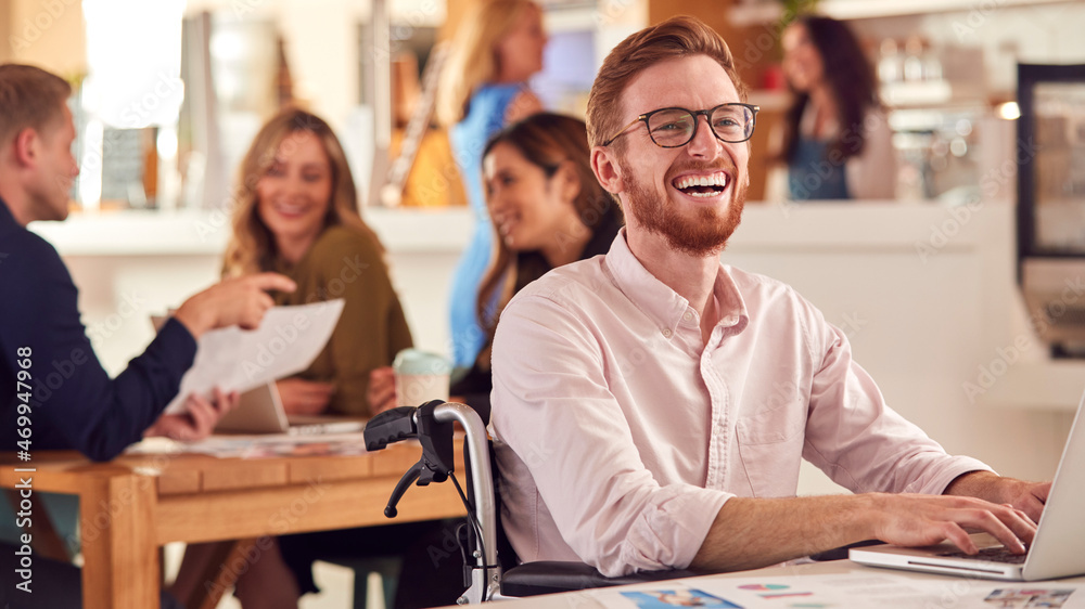 Laughing Businessman In Wheelchair Sitting At Desk Working On Laptop In Busy Office Laughing Businessman In Wheelchair Sitting At Desk Working On Laptop In Busy Office