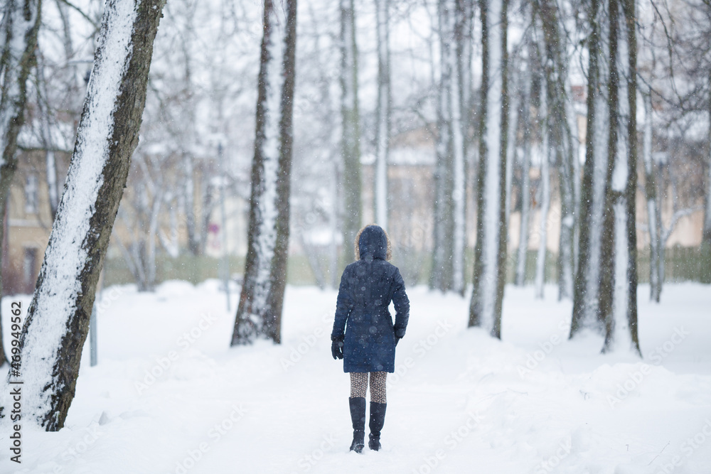 Young adult woman slowly walking through alley of trees in white snowy winter day at park. Fresh first snow. Spending time alone in nature. Peaceful atmosphere. Back view.