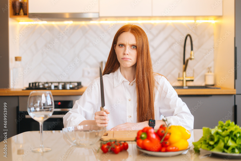 Portrait of attractive redhead young woman with angry eyes holding big knife sitting at table with cutting board and looking at camera in light kitchen room with modern interior, close-up. Portrait of attractive redhead young woman with angry eyes holding big knife sitting at table with cutting board and looking at camera in light kitchen room with modern interior, close-up.