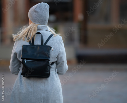 Happy young blonde teenage girl going to school or college wearing black backpack. Blonde girl in rgay hat and gray coat. Pictuce from back photo