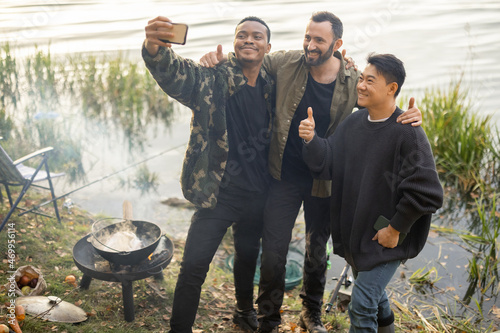 Smiling multiracial male friends taking selfie on smartphone during resting in nature. Men fishing and cooking Ukha soup on river coast at autumn. Leisure, weekend and vacation. Idea of friendship
