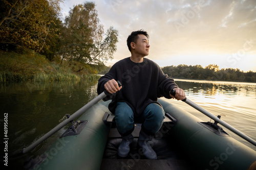 Asian man floating on rubber boat in lake or river at autumn morning. Concept of rest, weekend and vacation in nature. Idea of leisure outdoors. Adult male person wearing boots and warm clothes