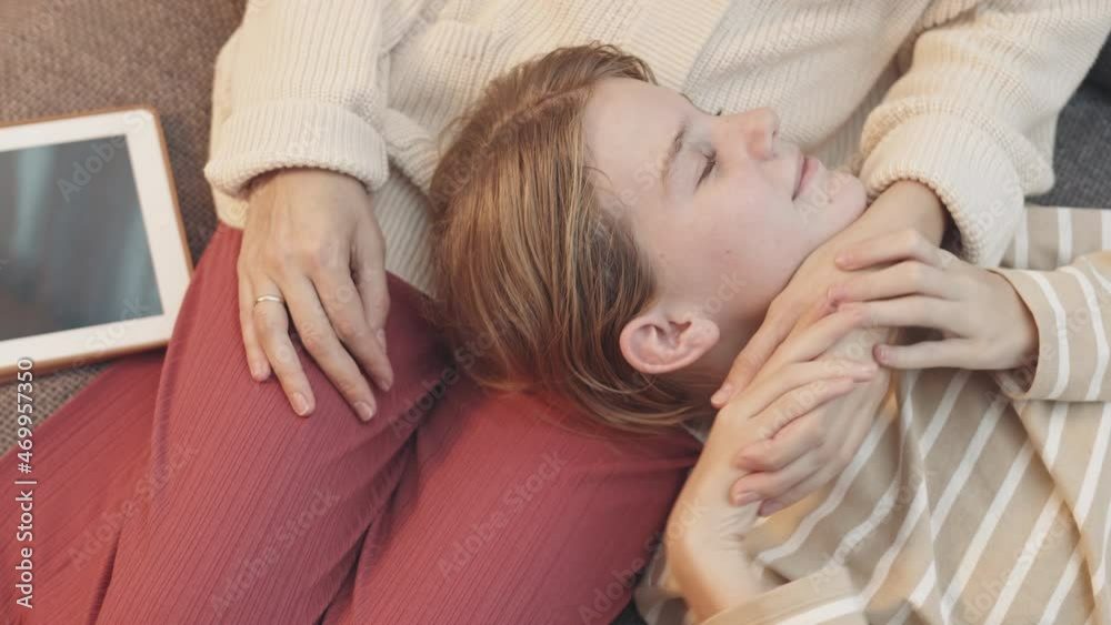 Top view of blue-eyed Caucasian teenage girl lying with head on lap of cropped mother, smiling and kissing her hand