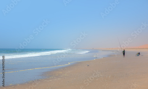 Fisherman fighting fish with a long fishing rod on Atlantic ocean beach in front of beautiful blue sky