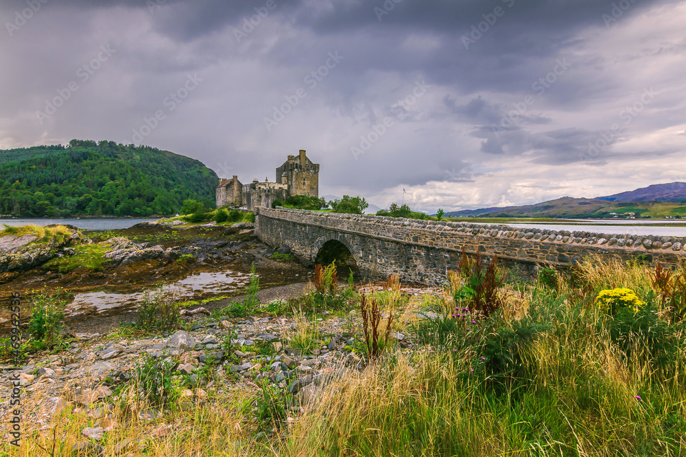 Eilean Donan Castle in Scotland with the historic stone bridge for pedestrians during the day when the water is low. Bushes and stones with algae in the foreground at low tide.