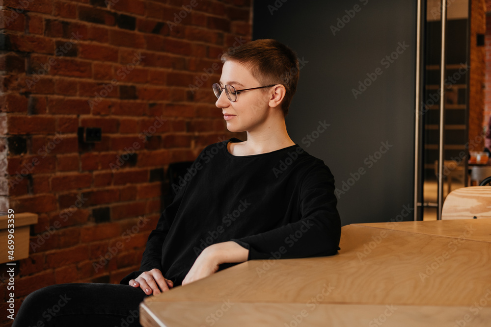 Smiling businesswoman with short hair in eyeglasses, black sweater sitting at the table. Confident young professional at work in the office. Successful freelacer in cafe Smiling businesswoman with short hair in eyeglasses, black sweater sitting at the table. Confident young professional at work in the office. Successful freelacer in cafe