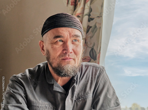 Portrait of senior asian muslim man with beard wearing skullcap sitting by window. photo