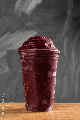 Brazilian Frozen Açai Berry Ice Cream Smoothie in plastic cup. On a wooden desk and a gray summer background. Front view for menu and social media
