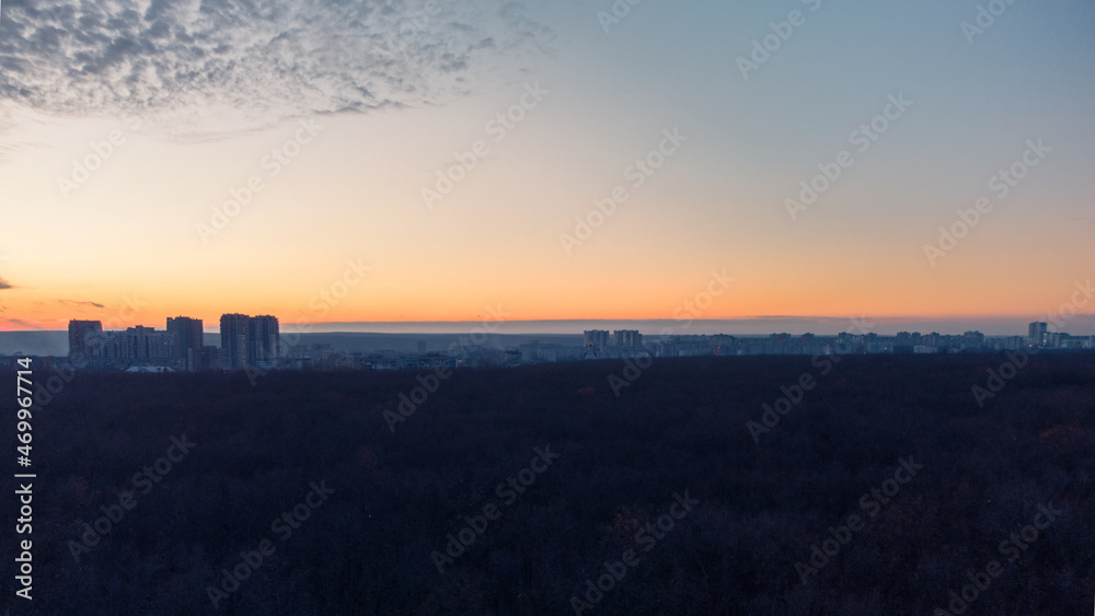 Aerial sunset evening wide view above dark blue autumn forest near residential Pavlove Pole district in Kharkiv city. Gray multistory buildings with scenic orange cloudy sky on horizon