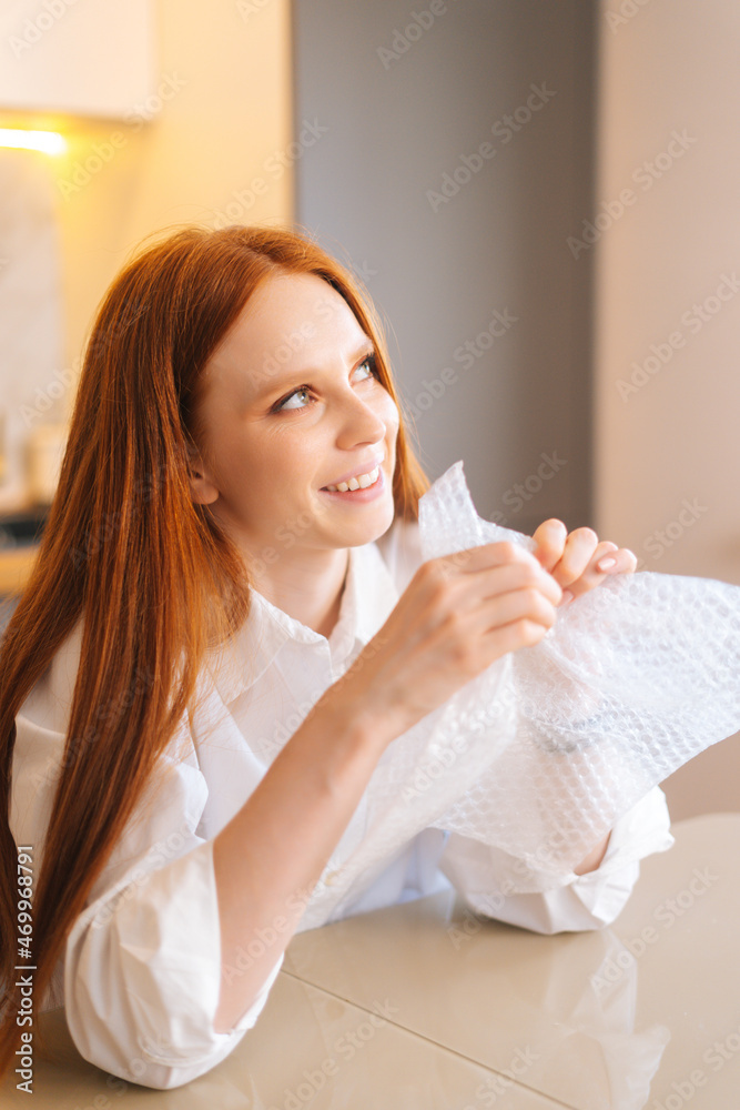 Close-up vertical shot of dreaming young woman playing pops bubble wrap to calm herself sitting at table in kitchen. Smiling pretty redhead female popping plastic bubblewrap. Concept of mental health.