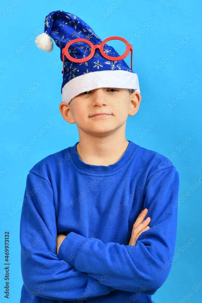 A funny boy in a blue Santa Claus hat and red glasses on his head crossed his arms and poses for New Year and Christmas. A funny boy in a blue Santa Claus hat and red glasses on his head crossed his arms and poses for New Year and Christmas.