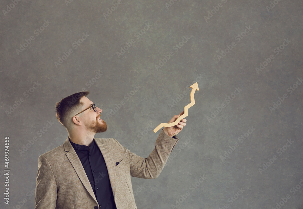 Good manager makes investment growth. Happy successful businessman holds a growth arrow and shows it up standing on a gray background. Business concept of growth, success and achievement. Good manager makes investment growth. Happy successful businessman holds a growth arrow and shows it up standing on a gray background. Business concept of growth, success and achievement.