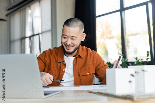 smiling asian architect holding smartphone while working with blueprints near blurred laptop photo
