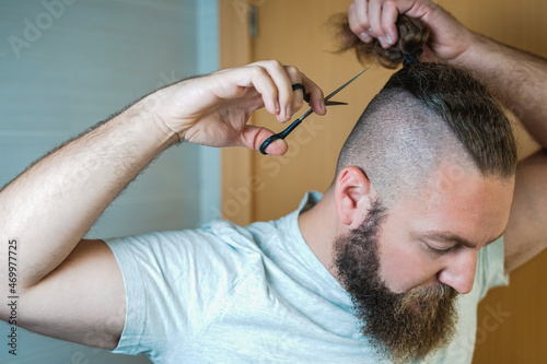 Caucasian man with long hair and beard cuts his ponytail with scissors.