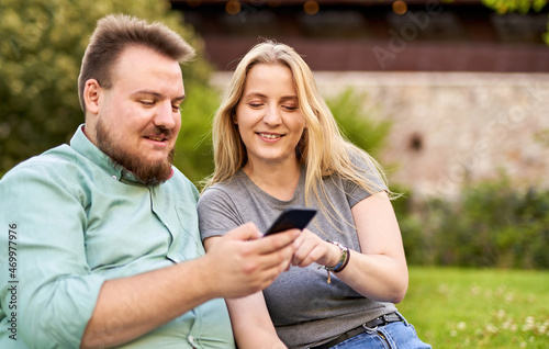 Young millennial couple checking social media and laughing together on a funny video in a park - Teenagers sharing a story on web - Modern technology and people lifestyle concept