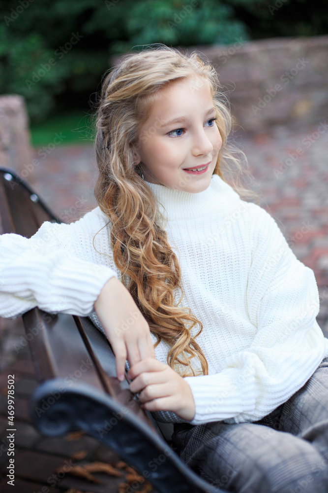 Obraz premium Blond girl on a bench in the park. Portrait of a blue-eyed girl in a white sweater in the park.