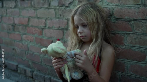 An unkempt little girl from a poor family plays with her toy against the backdrop of a brick wall.