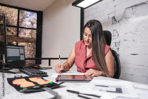 Hermosa mujer de negocios latina sentada en su oficina con un lápiz en su mano tomando apuntes de sus proyectos de arquitectura photo