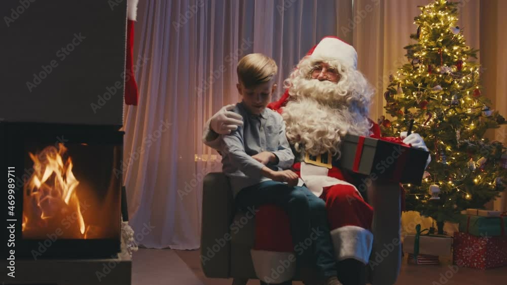 Little cute boy sitting near fireplace on Santa's knees, making a wish. Whitebearded Santa is hugging him and holding a gift.