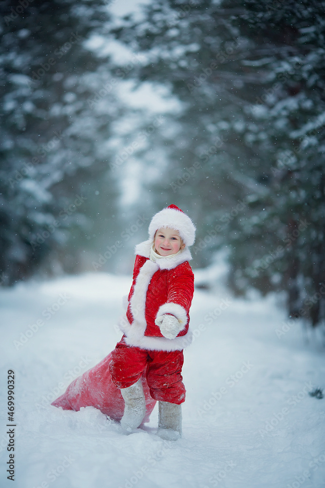 Little funny child dressed in Santa Claus red costume bringing presents in winter snowy forest. Christmas Eve.