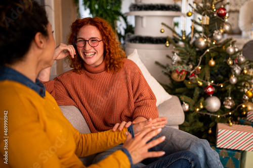 Two beautiful friends exchanging christmas gifts in the apartment smiling and laughing in front of a tree. Red haired and brunette enjoying new year's party. © bugarskipavle3
