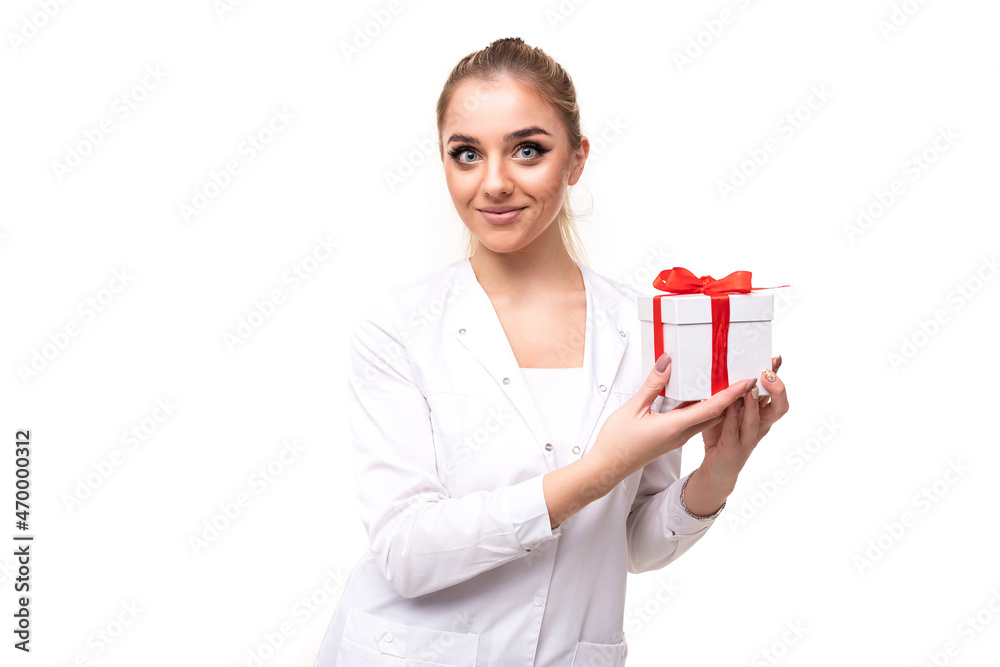 Photo of a beautiful pretty attractive positive smiling young blonde woman with a ponytail and makeup in a white medical coat of a nurse holding a white gift box with a red ribbon isolated on a white Photo of a beautiful pretty attractive positive smiling young blonde woman with a ponytail and makeup in a white medical coat of a nurse holding a white gift box with a red ribbon isolated on a white