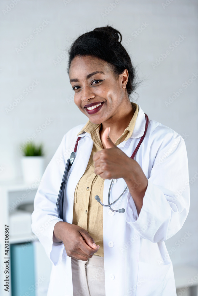 Female African American doctor wearing white coat with stethoscope standing in the office and looking at digital tablet Female African American doctor wearing white coat with stethoscope standing in the office and looking at digital tablet