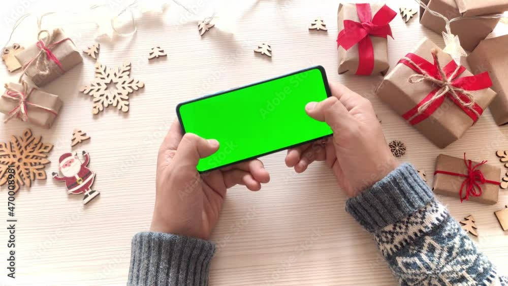 Man holding smartphone with horizontal green screen and touch on screen on Christmas background with gifts. Mobile Phone with Chroma Key. Close up hands. Man holding smartphone with horizontal green screen and touch on screen on Christmas background with gifts. Mobile Phone with Chroma Key. Close up hands.