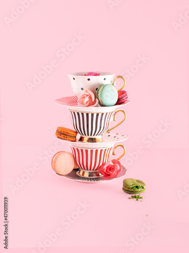 Colorful cups and saucers, creative composition, roses and macarons against pastel pink background. Tea time snack theme.